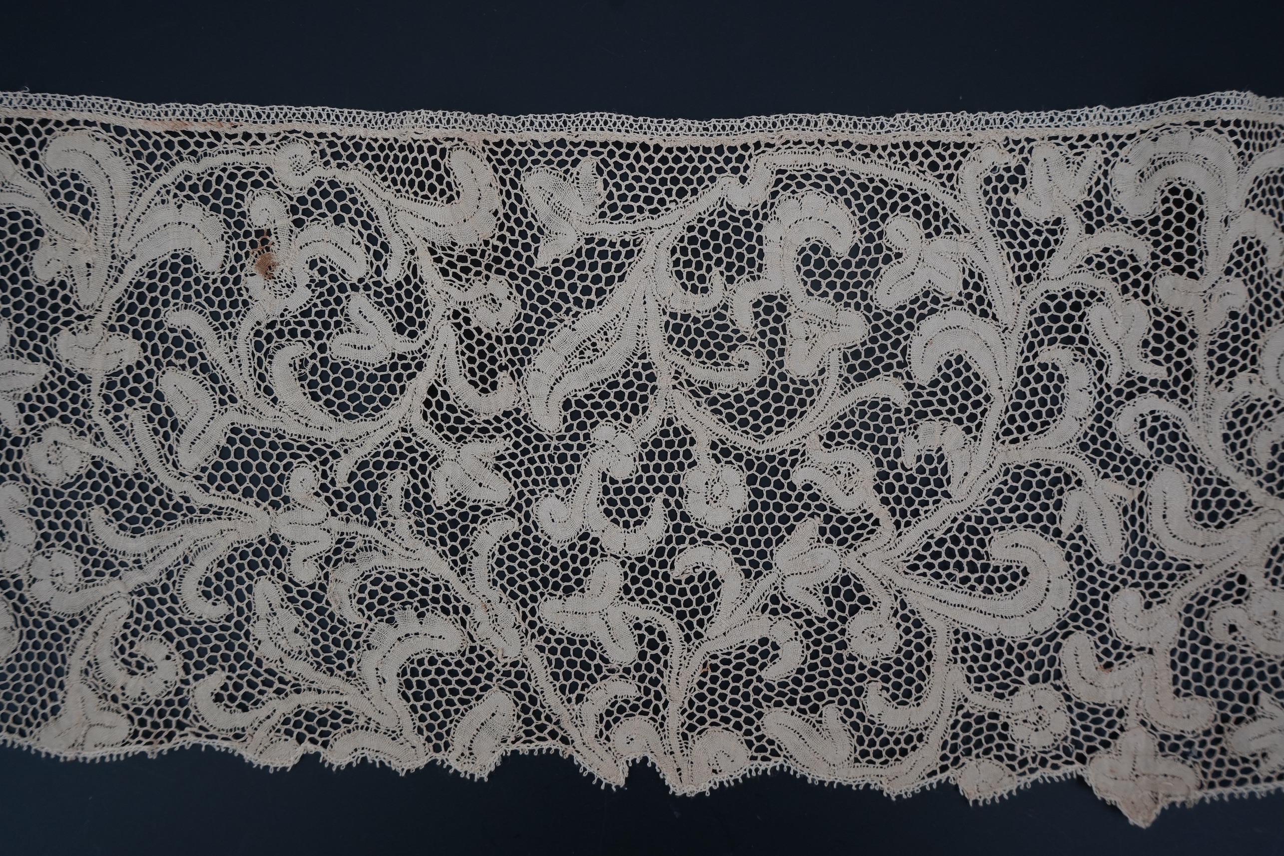 A length of 1740-1760 Italian, Milanese bobbin lace flounce, made from flat braiding or tapes, placed into scrolling patterns, attached to brides, made into a honeycomb mesh. 252cm long x 16cm deep. Condition - one small